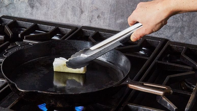 A hand holding a greased paper towel with tongs to season a cast iron skillet on the stove.