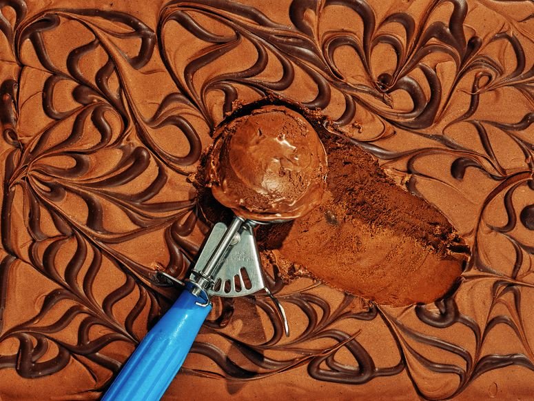A blue ice cream scoop holding a scoop of chocolate ice cream atop the metal pan of ice cream.